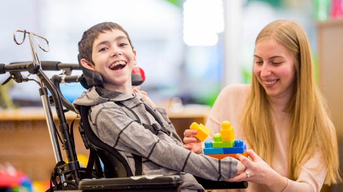 Young boy suffering from cerebral palsy laughing on a wheelchair with his mother nearby