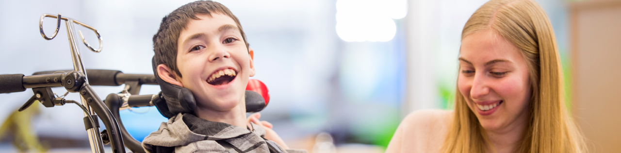 Young boy suffering from cerebral palsy laughing on a wheelchair with his mother nearby