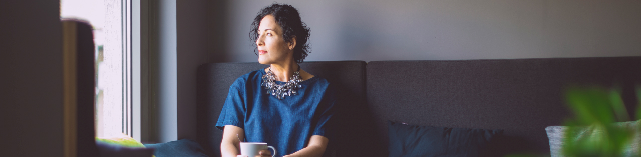 Woman sitting on sofa with mug of tea looking outside sadly after having suffered from sexual abuse