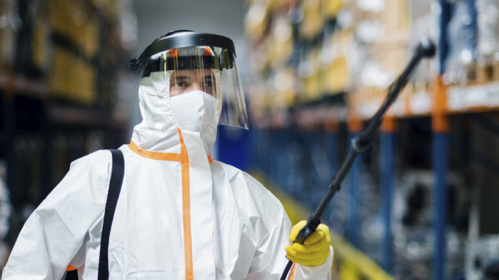 Person in white hazmat suit with face covering and yellow gloves holding specialist equipment to remove industrial disease from warehouse