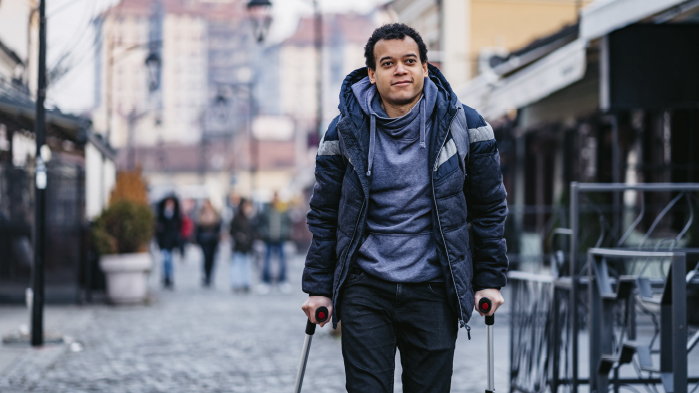 Young disabled man with ctruches who suffered personal injury is standing outside on the street wearing hoodie jumper and coat